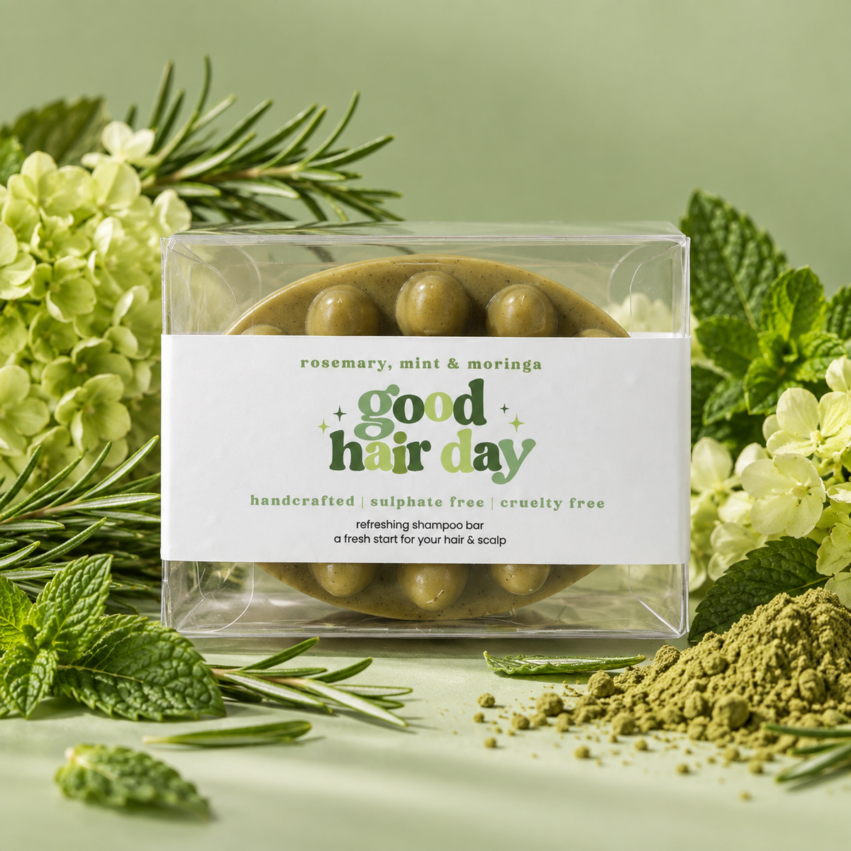 Good Hair Day rosemary, mint and moringa shampoo bar in a clear box, surrounded by herbs and green powder.
