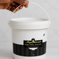 White bucket of Shea Nilotica with a black label held by a hand on a light grey background