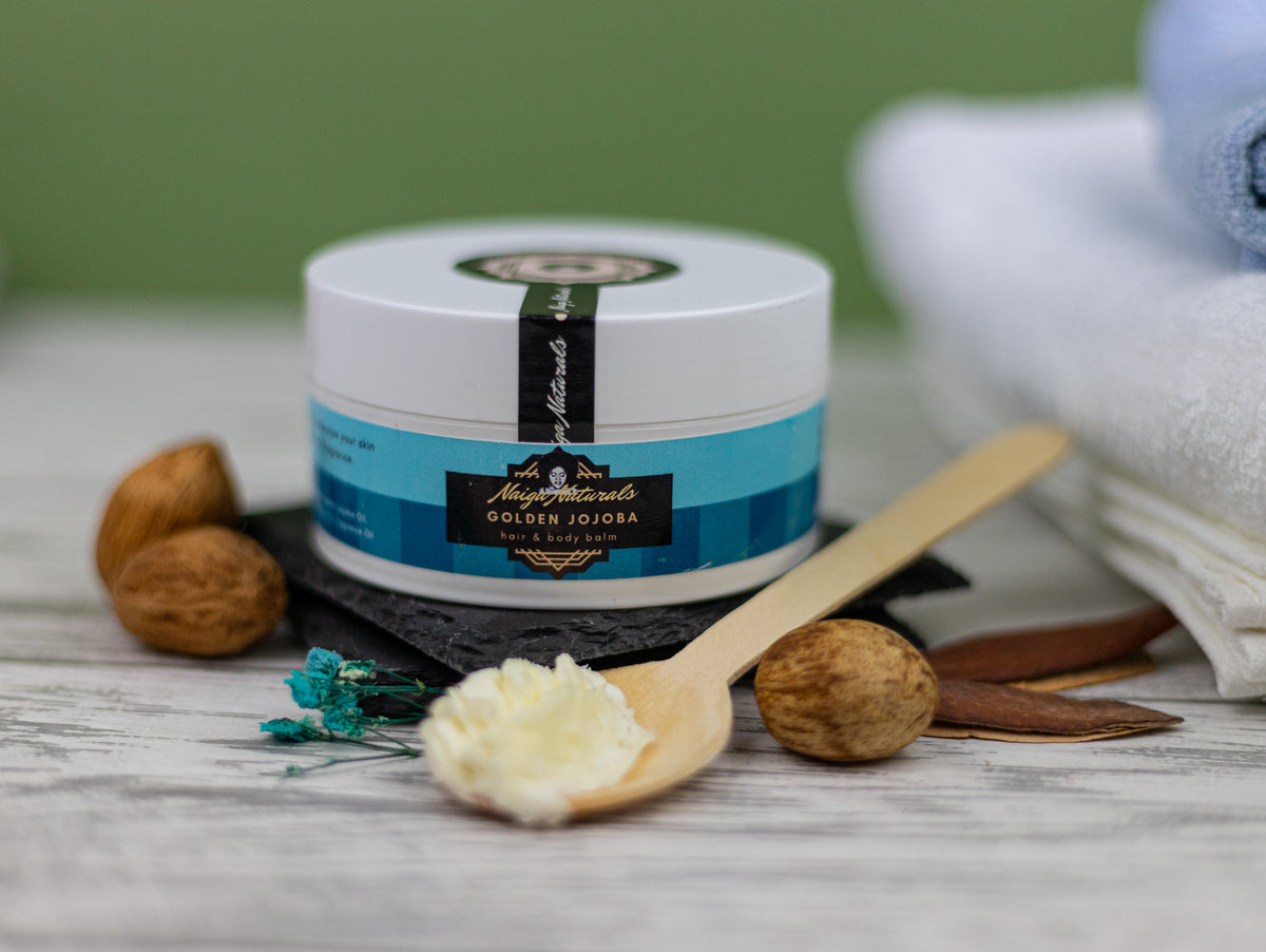 Naiga Naturals Golden Jojoba Hair & Body Balm jar with a blue label beside a wooden spoon holding pale balm.
