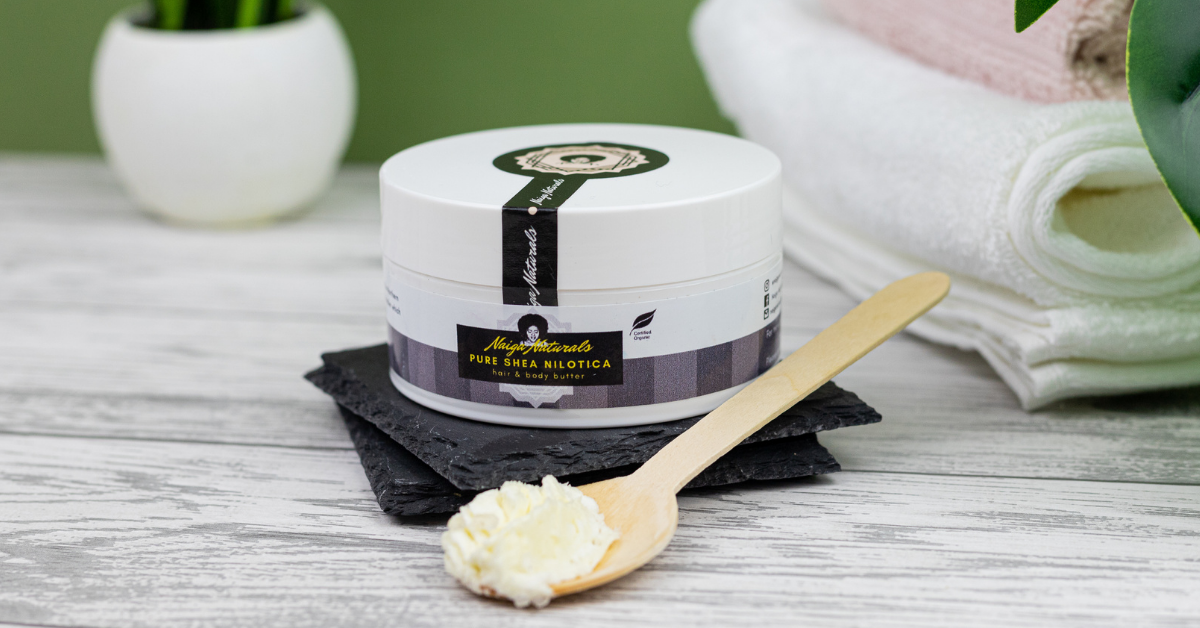 Naiga Naturals Pure Shea Nilotica butter jar on black slate beside a wooden spoon holding pale shea butter.
