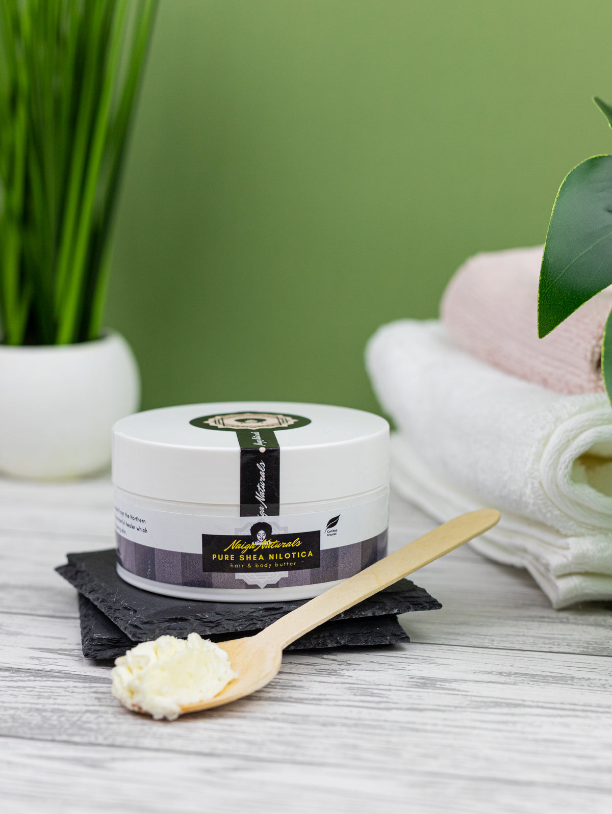 Naiga Naturals Pure Shea Nilotica butter jar and a spoonful of shea butter beside folded towels and a potted plant.