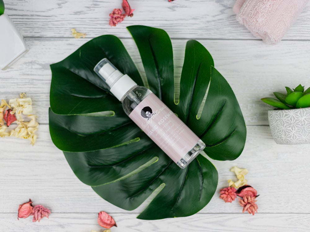 Naiga Naturals Rosewater Mist with Glycerine spray bottle resting on a green leaf, surrounded by dried rose petals.
