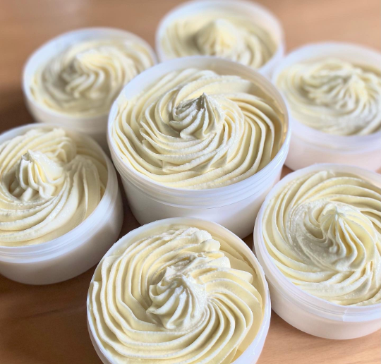 Open white tubs filled with pale whipped hair and body butter piped into swirls on a wooden surface.
